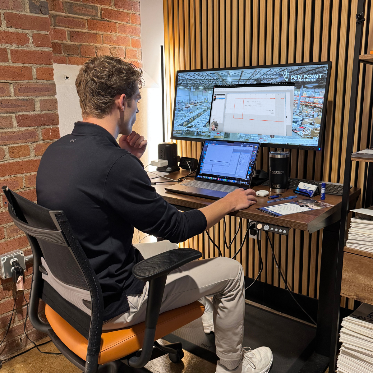 A Pen Point support team member working at a multi-monitor desk with the Louisville warehouse feed on screen