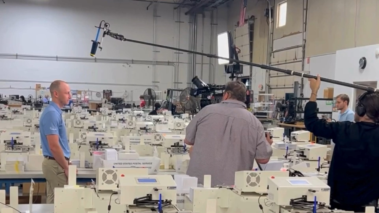 Matt Redmon being filmed on the Pen Point production floor, rows of handwriting machines running behind him